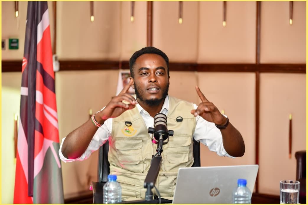 Denzel Maina speaking at a microphone behind a desk with a laptop, a Kenyan flag to one side