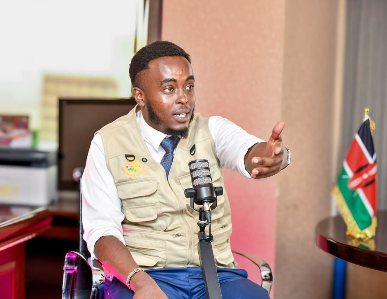 Denzel Maina gesturing as he speaks into a microphone, a Kenyan flag beside him