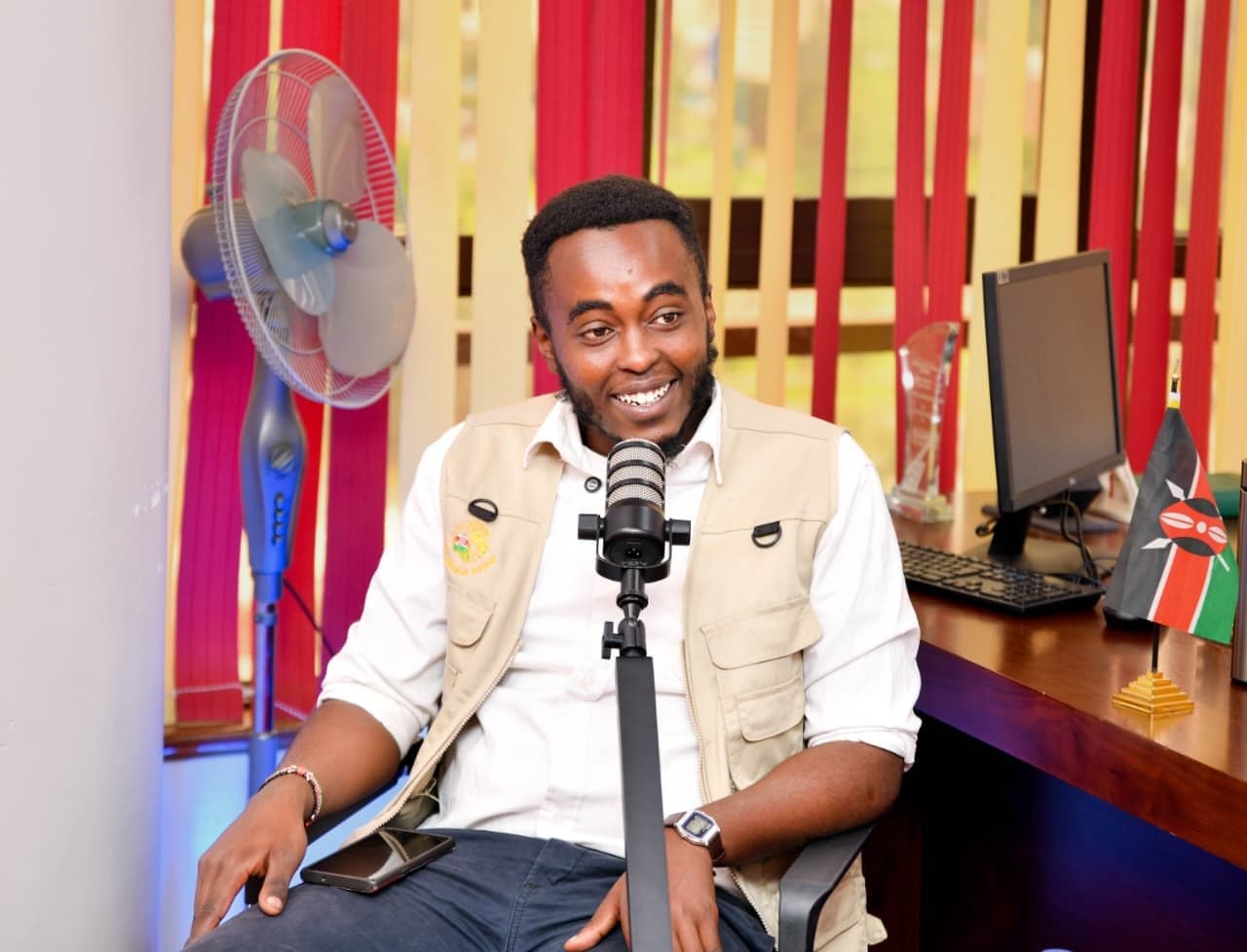 Denzel Maina smiling at a podcast microphone in an office, a Kenyan flag on the desk behind him